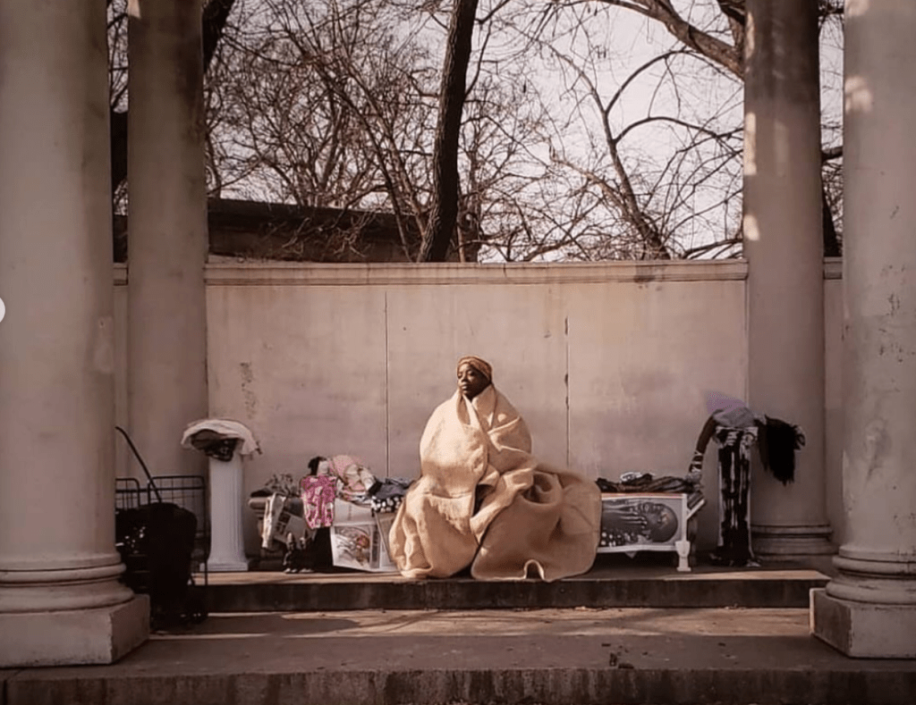 A photograph of a Black woman draped in a beige blanket sitting on a bench in a public space with neoclassical columns.