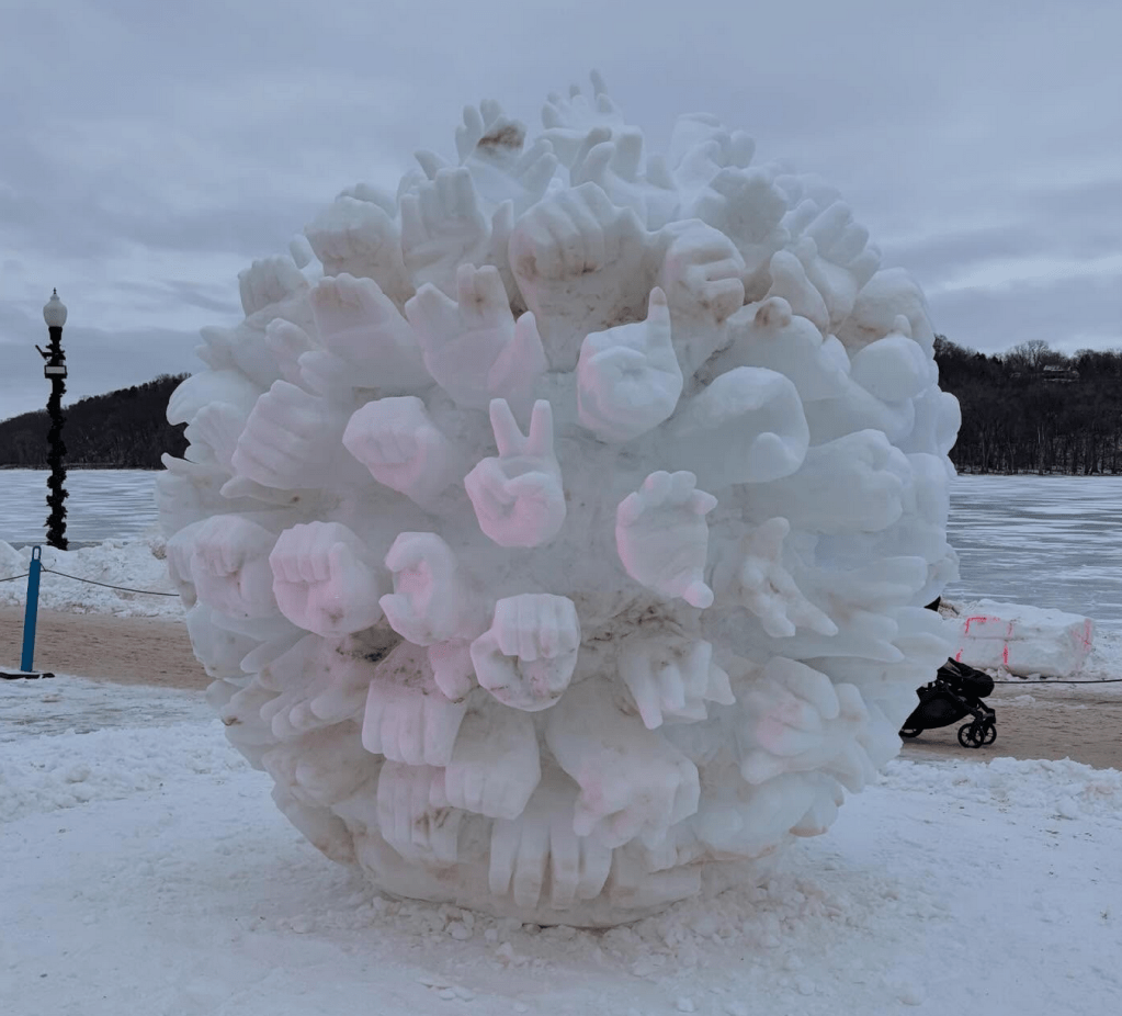 A large spherical ice sculpture in a park with protruding hands signing words and phrases in ALS, such as “ICE out,” “love,” “unity” and “resist.”