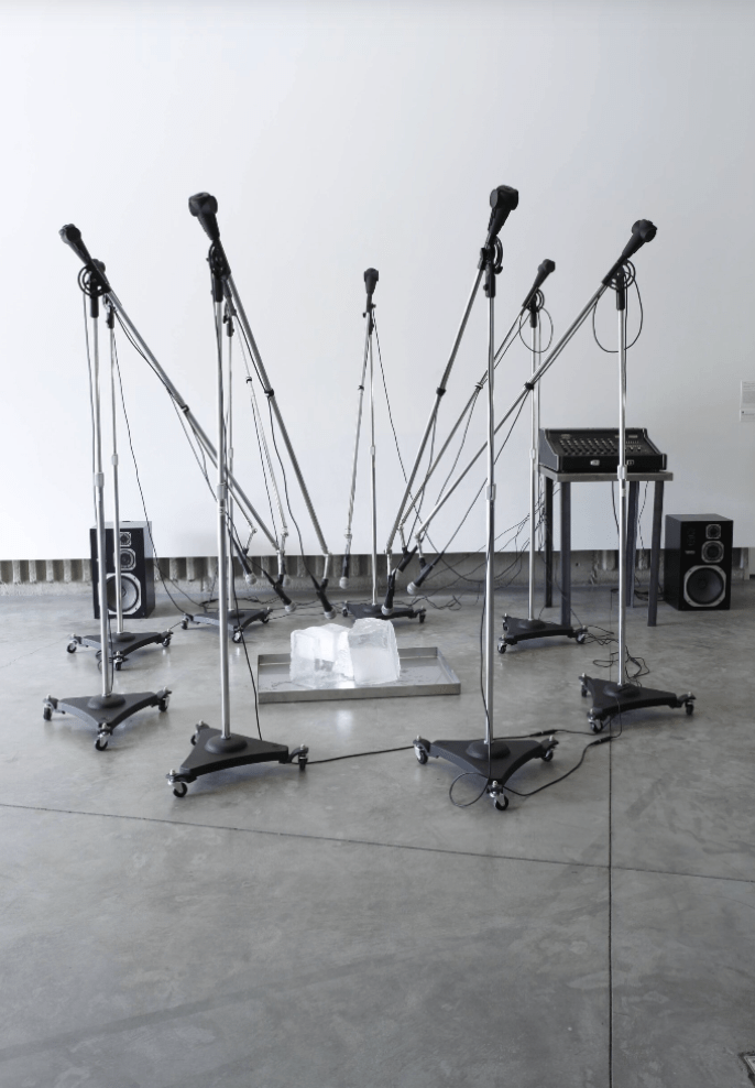 A conceptual art installation of eight boom microphones pointing downward to a tray of large ice blocks in the center. 