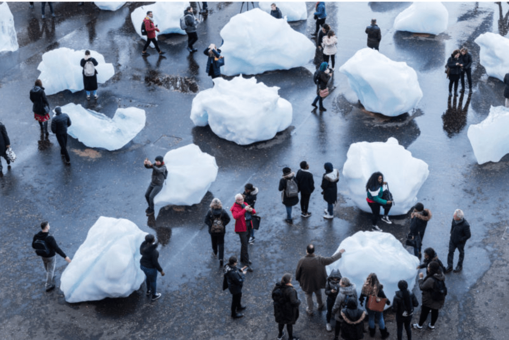 Aerial view of several large blocks of ice arranged in a public plaza as an art installation. Groups of people in coats walk around the ice, some touching and leaning on it. 