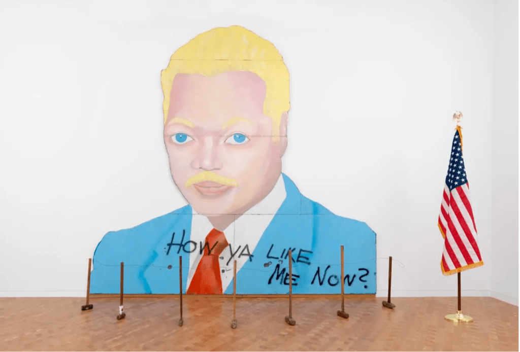 A large mural sized painting of Jesse Jackson reimagined as a white man with blonde hair, facial hair and blue eyes, wearing a blue suit and red tie. The words "How ya like me now?" are spray painted across his chest. In front of the painting is a stanchion type of barrier made of sledgehammers, with an American flag on a pole to the right hand side.