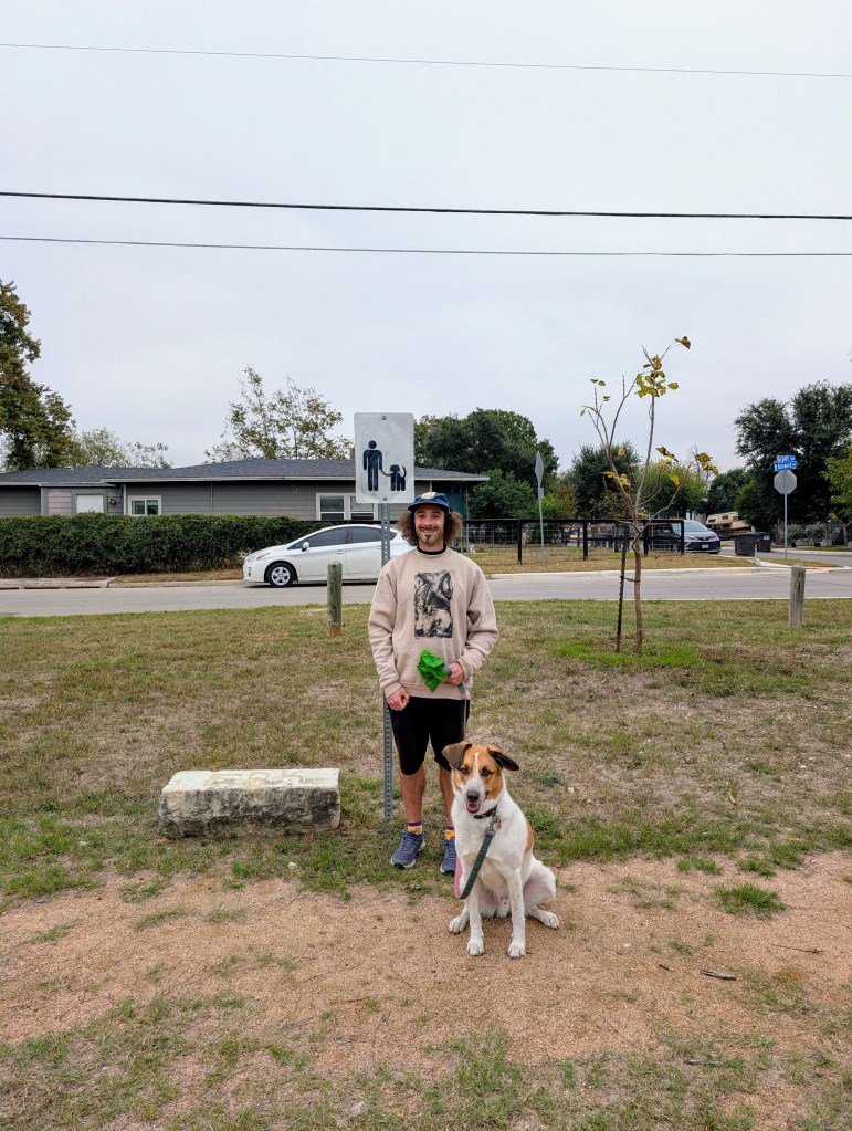 A photograph of a man in a beige wolf sweatshirt holding a leash with a beige and white, large shepherd mix sitting down in front of him. The man and dog are in a local park, standing behind a sign with icons of a human and dog on a leash. 
