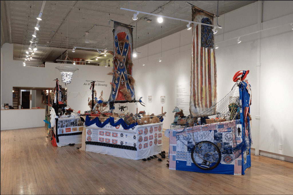 A sculpture of a vessel, broken into three parts. The front of the ship has a masthead featuring Uncle Sam and represents large corporations' influence on American society; another features imagery depicting class struggle and racial disparity; and a final section include symbols depicting the social elite. 